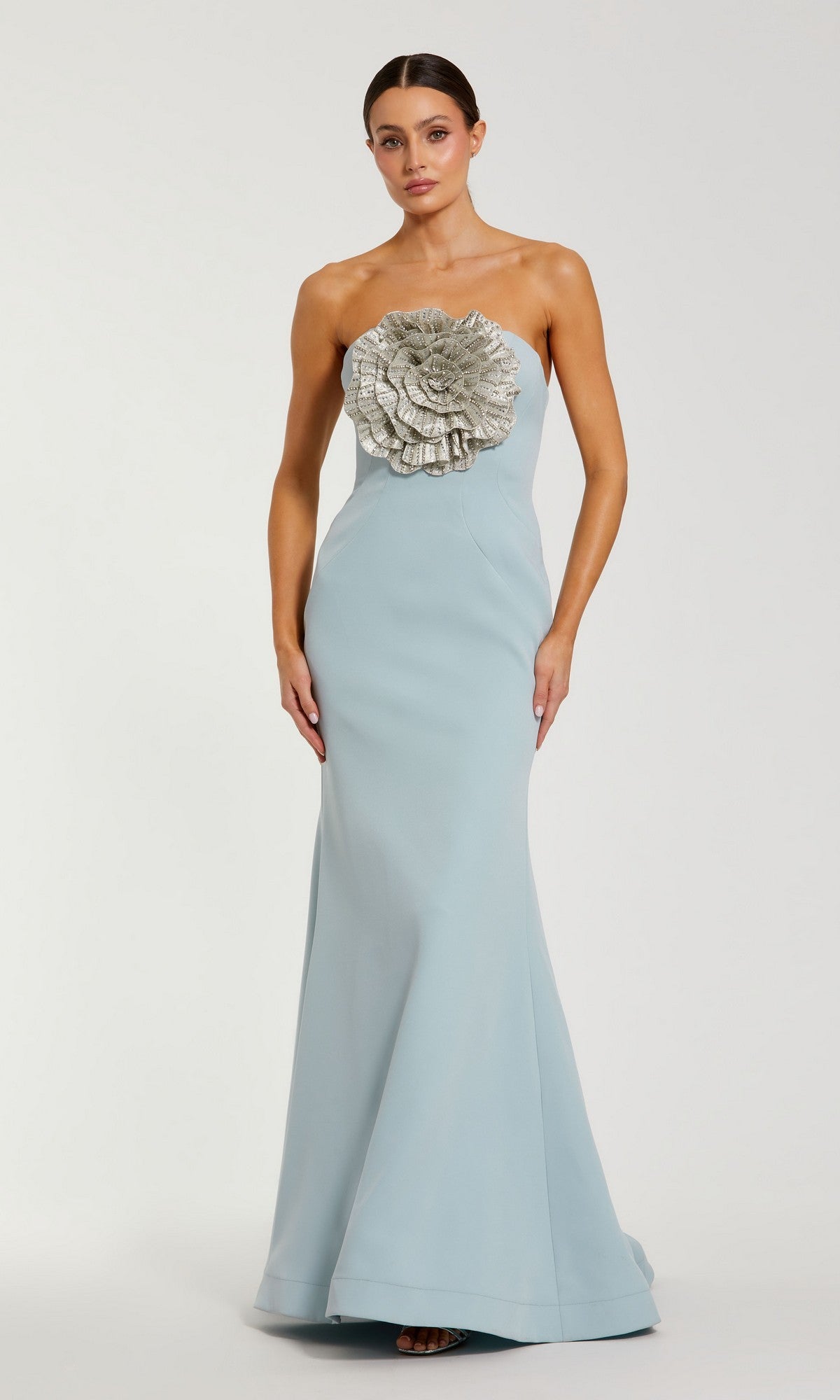 A woman models the Mac Duggal 11855 Strapless Floral Formal Gown in powder blue, featuring a long silhouette and a large silver ruffled floral embellishment on the bodice, set against a simple pale background.